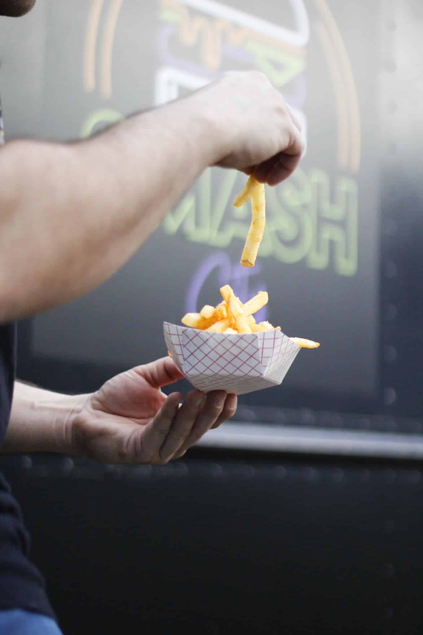 A person holding a paper bowl of fries, picking one up, with a colorful truck logo blurred in the background.