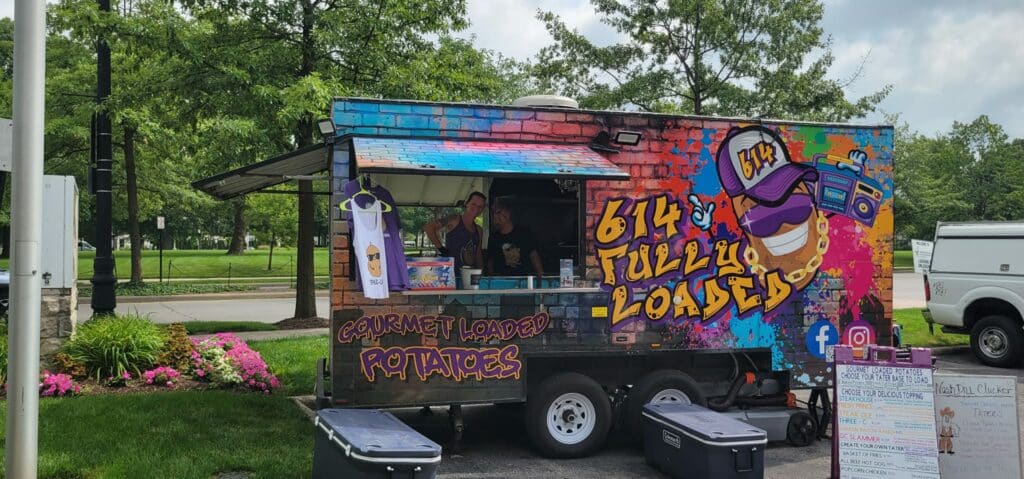 Colorful food truck in a park selling gourmet loaded potatoes, with a vibrant graffiti-style design featuring a skull, and two staff members visible at the service window.