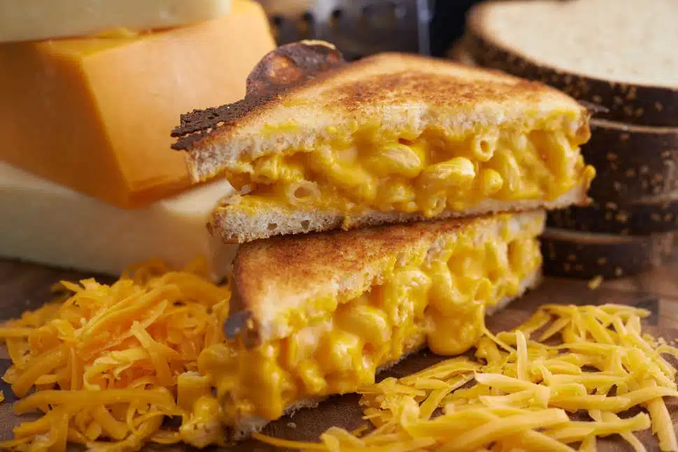 A grilled cheese sandwich with melted macaroni and cheese filling, cut in half, on a wooden board surrounded by shredded cheese.