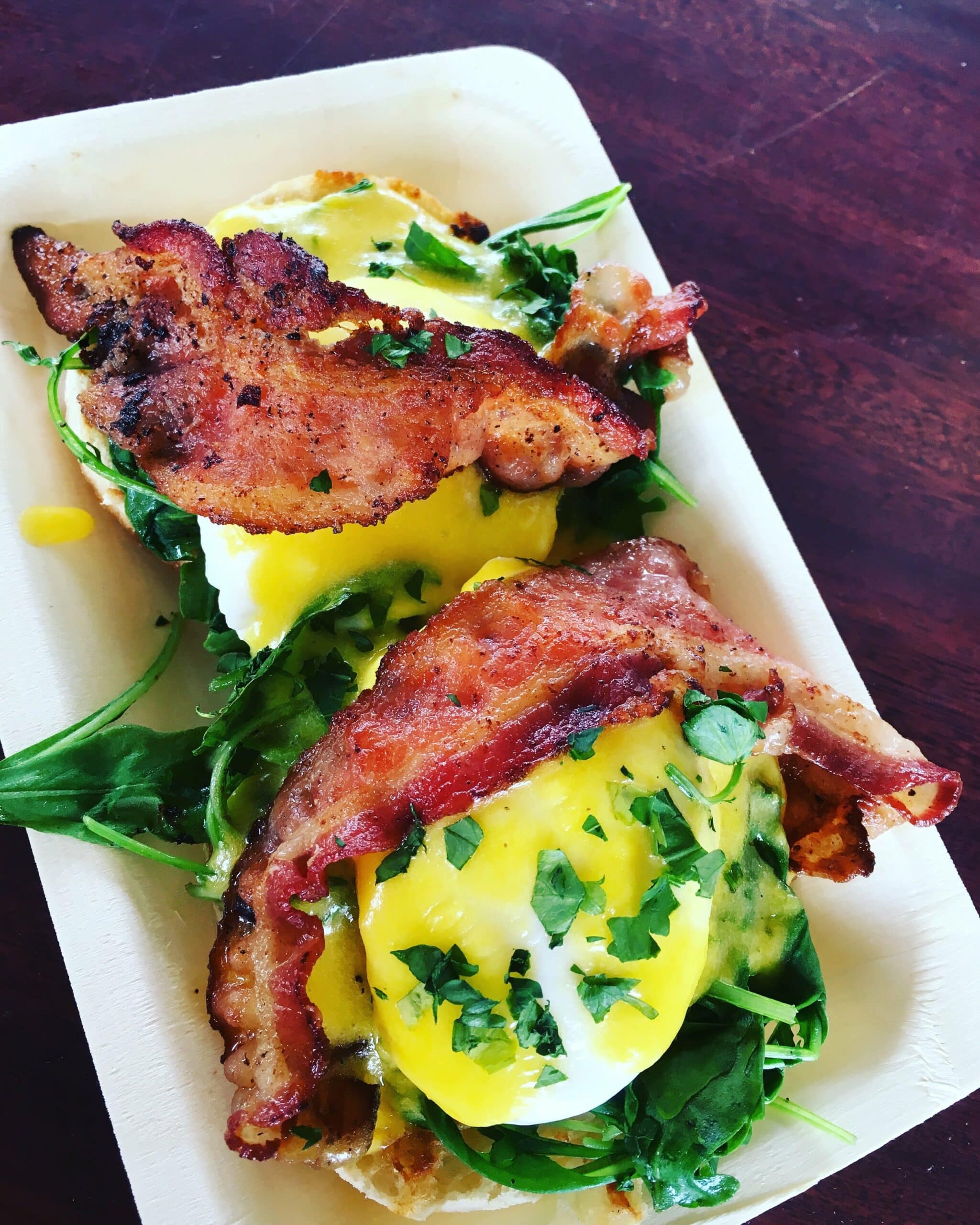 A dish featuring eggs benedict with hollandaise sauce, bacon, and greens served on a rectangular, biodegradable plate.