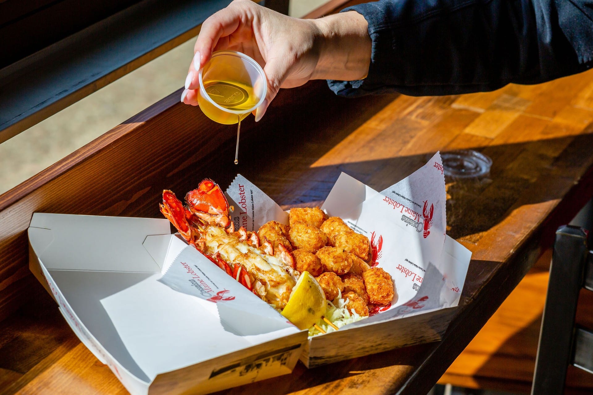 A person pours melted butter over a meal consisting of lobster, fried shrimp, and a lemon wedge, served in a white takeout container on a wooden table.
