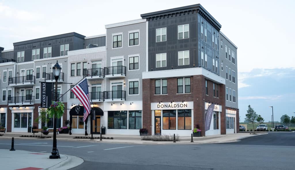 A modern multi-story building in the Evans Farm neighborhood features businesses on the ground floor, including "Donaldson." An American flag proudly waves in front of the building, with a streetlamp nearby.