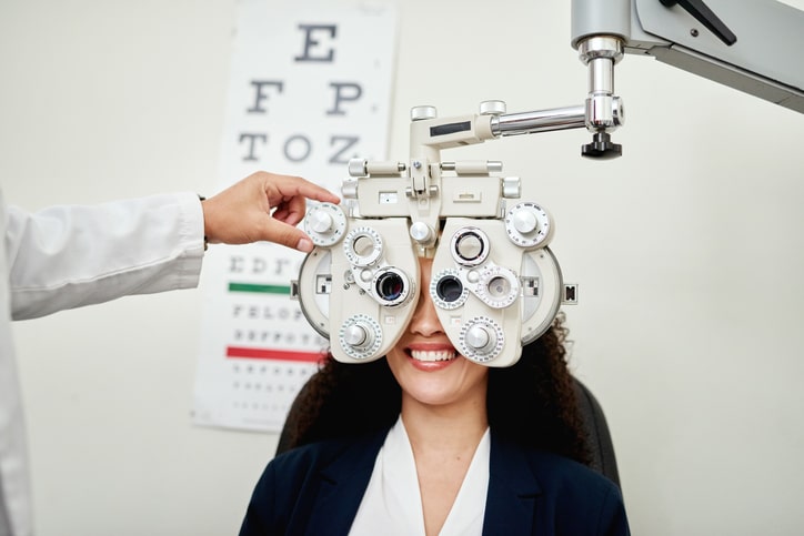 A woman undergoing an eye examination with a phoropter, while the optometrist adjusts the device in their Evans Farm neighborhood office. An eye chart is visible in the background.