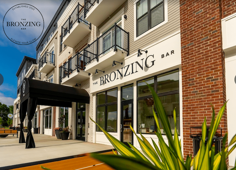 Street view of The Bronzing Bar in the Evans Farm neighborhood, showcasing a black awning and modern façade with balconies and large windows, all surrounded by lush greenery.