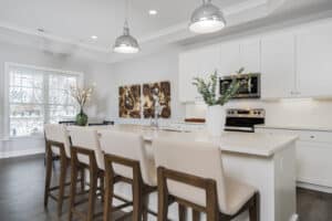 Modern kitchen with white cabinets, stainless steel appliances, island with four beige chairs, pendant lights, and decorative vases with greenery. Large window provides natural light.