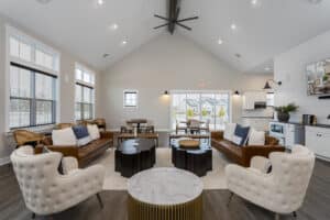 Modern living room with vaulted ceiling, brown leather sofas, white tufted chairs, coffee tables, large windows, and an open kitchen in the background.