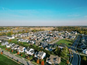 Aerial view of a suburban neighborhood with rows of houses, green lawns, streets, and trees displaying autumn colors under a clear blue sky.