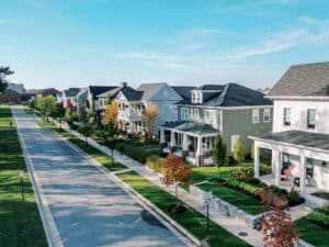 Aerial view of a suburban street lined with modern houses, trees, and a sidewalk. A 25 mph speed limit sign is visible. The sky is clear and blue.