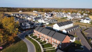 Aerial view of a suburban neighborhood with a curved road, brick townhouses in the foreground, and detached houses in the background surrounded by fall foliage.