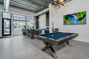 Modern loft apartment interior with a pool table in the center, a kitchen area with bar stools in the background, and a colorful abstract painting on the wall.