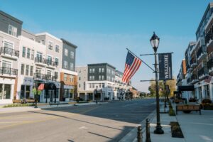 A quiet, modern street with multi-story apartment buildings, an American flag, a "Welcome" banner, and empty sidewalks under a clear blue sky.