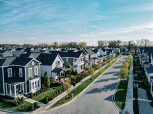 Aerial view of a suburban neighborhood with rows of modern houses, green lawns, and a tree-lined street under a clear blue sky.