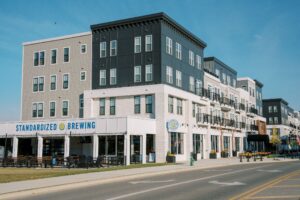 A modern, multi-story mixed-use building with "Standardized Brewing" signage, outdoor seating, and apartments above, situated along a wide street.
