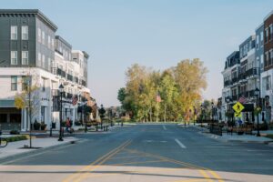 A wide, empty street runs through a modern suburban neighborhood with apartment buildings, trees, and American flags lining the sidewalks under a clear sky.