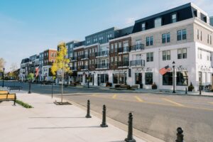 Modern apartment buildings with ground-floor shops line a quiet street, featuring American flags, young trees, and clear skies.