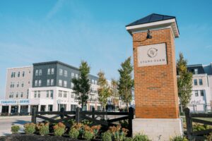 A brick monument with "Evans Farm" stands in front of modern apartment buildings and shops on a sunny day.