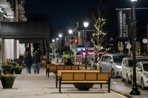 A city sidewalk at night with benches, planters, lit trees, parked cars, and a few people walking; streetlights and welcome banners are visible.