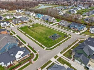 Aerial view of a suburban neighborhood with houses surrounding a central grassy park area that includes a playground. Roads border the park and houses line the streets.