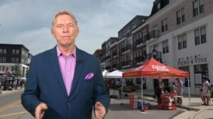 An older man in a blue suit stands in front of a street market with a red tent and apartment buildings in the background.