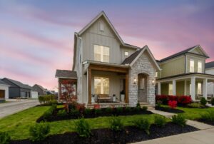 Two-story modern house with stone and siding exterior, front porch, lit windows, landscaped yard, and neighboring homes at sunset.