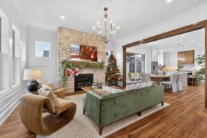 A modern living room with a stone fireplace, Christmas decorations, a green sofa, two brown chairs, and an open view into a kitchen and dining area.