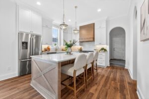 A modern kitchen with white cabinets, stainless steel appliances, a center island with three upholstered chairs, pendant lights, and wood flooring.