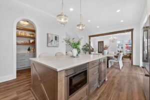 Modern kitchen with a large island, built-in microwave, pendant lights, open shelving pantry, and a view into a dining area with a table and chairs.