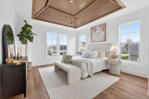 Modern bedroom with a white bed, bench, and rug, wood ceiling detail, large windows, plants, a black dresser with decor, and neutral-toned artwork on the wall.
