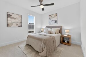 A modern bedroom with a neatly made bed, two abstract paintings on white walls, a wooden nightstand with lamps, and a ceiling fan. Natural light enters through a single window.