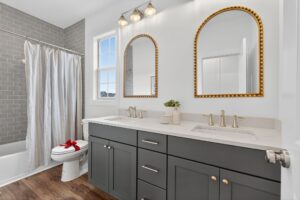 Modern bathroom with double sink vanity, two gold-framed mirrors, gray cabinets, a shower with gray tile, and a toilet with a red bow on the lid.