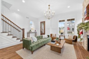 Bright living room with a green sofa, two tan chairs, a wooden coffee table, and a chandelier. Stairs and a decorated fireplace are visible, along with windows showing houses outside.