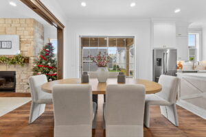 A modern dining area with a round wooden table, six upholstered chairs, a vase with branches, and a view of a decorated living room and backyard through large windows.