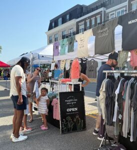 People browse t-shirts at an outdoor market stall on a sunny day, with apartment buildings visible in the background.