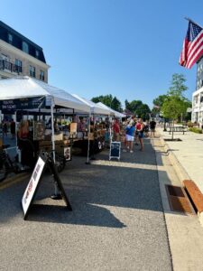People browse vendor tents at an outdoor market on a sunny day; a U.S. flag is visible, and a sandwich board sign stands in the foreground.