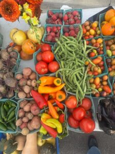 Assorted fresh vegetables, including tomatoes, green beans, potatoes, peppers, radishes, and eggplants, displayed in baskets at a market stall.