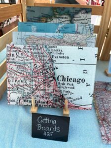 Cutting boards featuring a vintage map of Chicago and surrounding areas are displayed on a wooden stand with a sign reading "Cutting Boards $25".