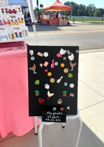 A display board with assorted handmade magnets is shown on a small table at an outdoor market. A sign reads "Magnets $7 each, 2 for $12.