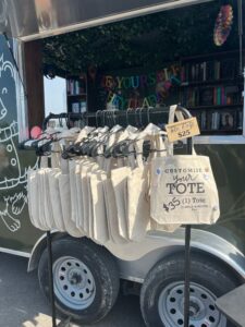 A rack of beige tote bags for sale in front of a trailer, with a sign reading “Customize Your Tote $25 (1) Tote.”.
