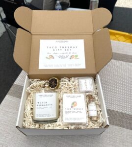 An open box labeled "Taco Tuesday Gift Set" containing a candle, two small jars, and a small bottle, all packed with shredded paper.