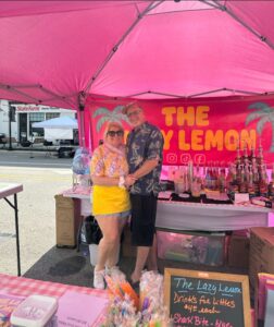 Two people stand smiling together under a bright pink tent labeled "The Lazy Lemon" at an outdoor market, surrounded by drink ingredients and a menu board.