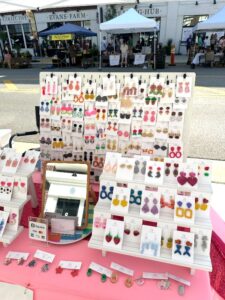 A jewelry display at an outdoor market featuring various colorful handmade earrings and a mirror on a pink tablecloth, with shops and people visible in the background.