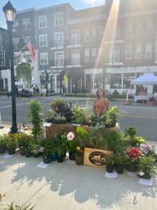A woman stands behind a table with potted plants and flowers for sale on a sunny sidewalk in front of shops and apartments.