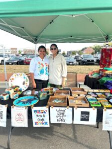 Two people stand behind a table displaying colorful arts and crafts items under a green canopy at an outdoor market.