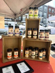 Bottles of spice blends are displayed on stacked wooden crates at an outdoor market booth, with buildings visible in the background.