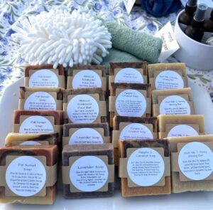 A display of handmade soap bars labeled with different scents and ingredients, arranged neatly on a table with washcloths and spray bottles in the background.