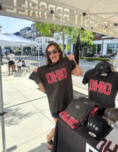 A woman stands under a tent at an outdoor market, smiling and holding up a black T-shirt with "OHIO" in large red letters. Other Ohio-themed merchandise is displayed on the table.