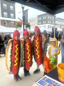 Four children in costume stand outdoors at an event; three are dressed as hot dogs, and one is dressed as a yellow animal, possibly a duck or chick, holding treat bags.