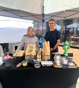 Two vendors stand behind a market stall displaying bread, pastries, and condiments under a canopy at an outdoor market.