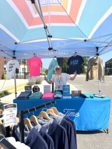 A person stands at an outdoor merchandise booth under a canopy, displaying T-shirts, hats, and sweatshirts for sale.