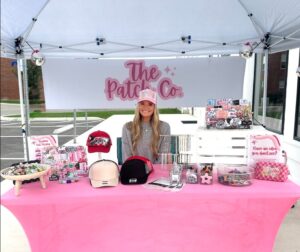 A woman sits behind a pink table displaying hats, patches, and accessories under a tent with a sign reading "The Patch Co.